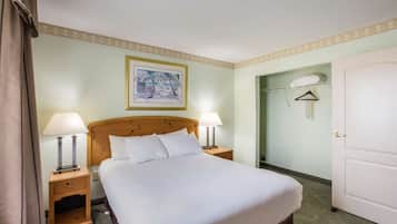 A neatly made bed with white linens, two bedside lamps, a wooden nightstand, a framed picture on the wall, and a door to a bathroom.