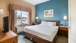 Une chambre d’hôtel avec un grand lit, deux tables de chevet avec des lampes, un téléviseur posé sur un bureau en bois, une fenêtre avec des rideaux et un tableau accroché au mur.