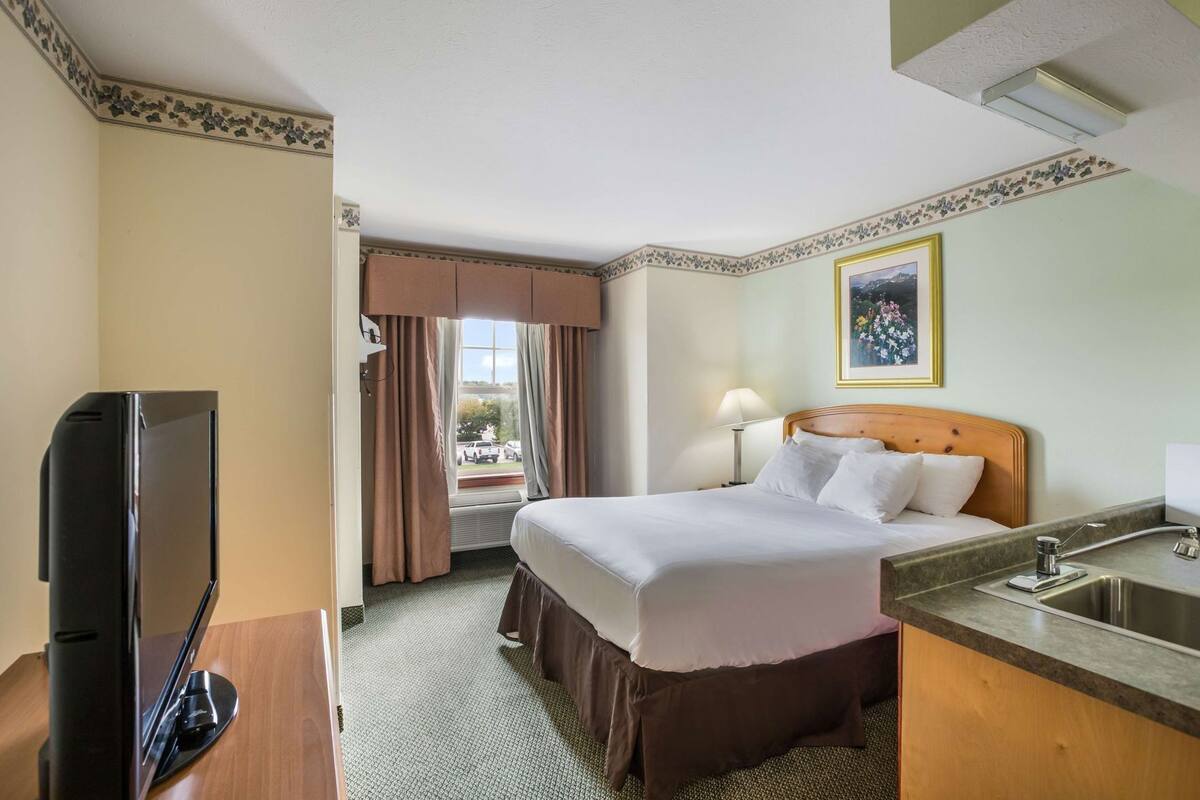 A hotel room with a bed, a TV, a desk with a sink, and a window with curtains.