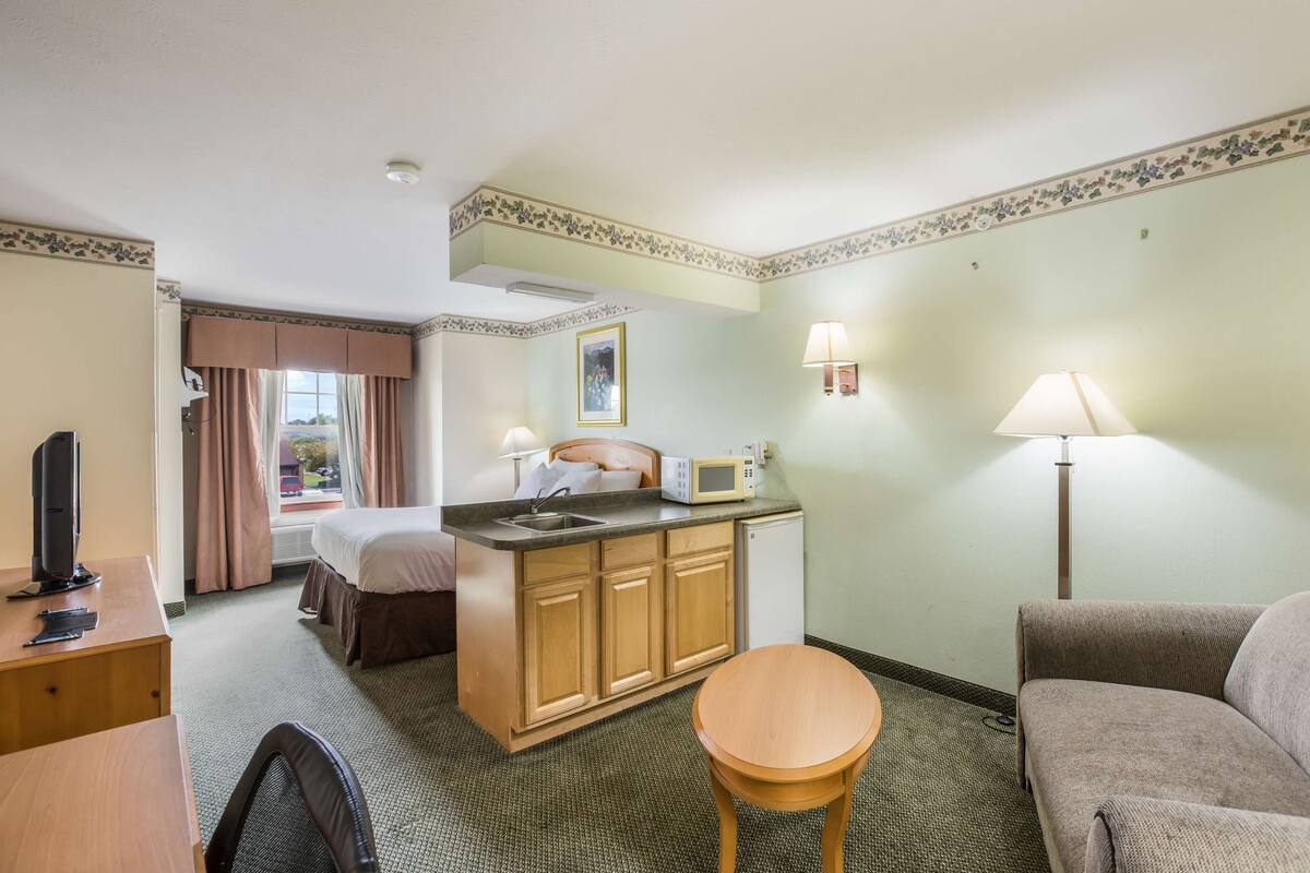 A hotel room with a bed, a desk, a small kitchenette, a sofa, and a lamp.