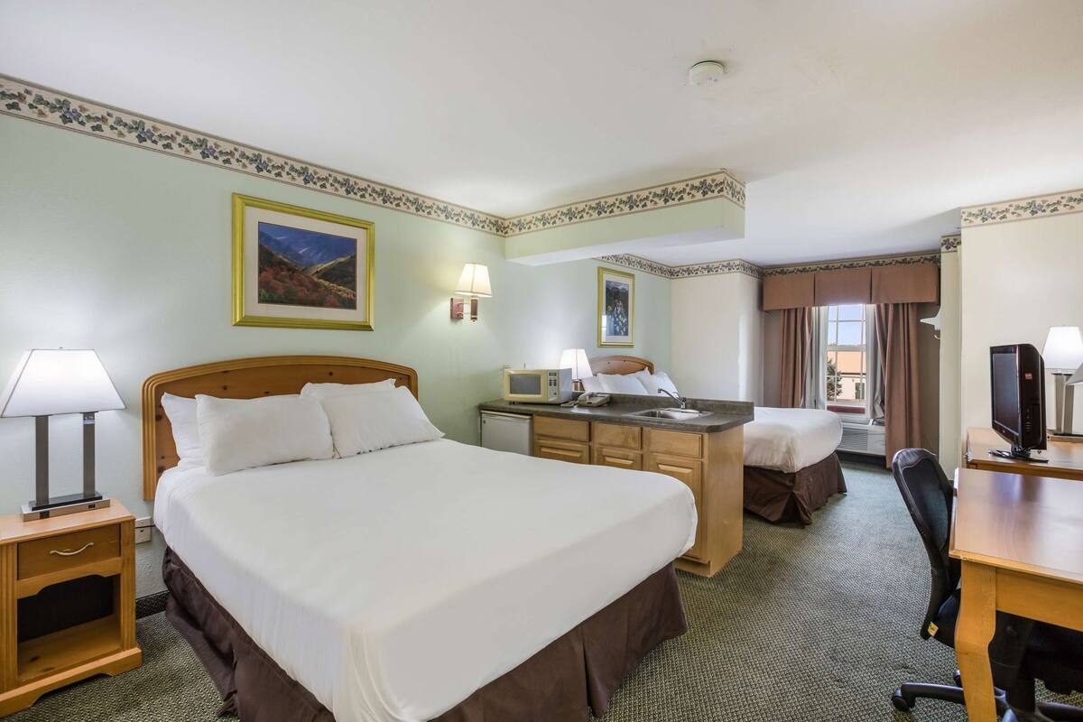 A hotel room with a large bed, a desk with a computer, a small kitchenette, and a window with curtains.