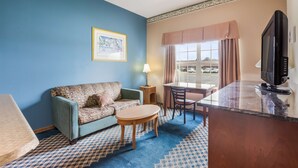Une chambre d’hôtel avec un mur bleu, un canapé à motifs, une petite table ronde, un téléviseur et une fenêtre avec des rideaux.
