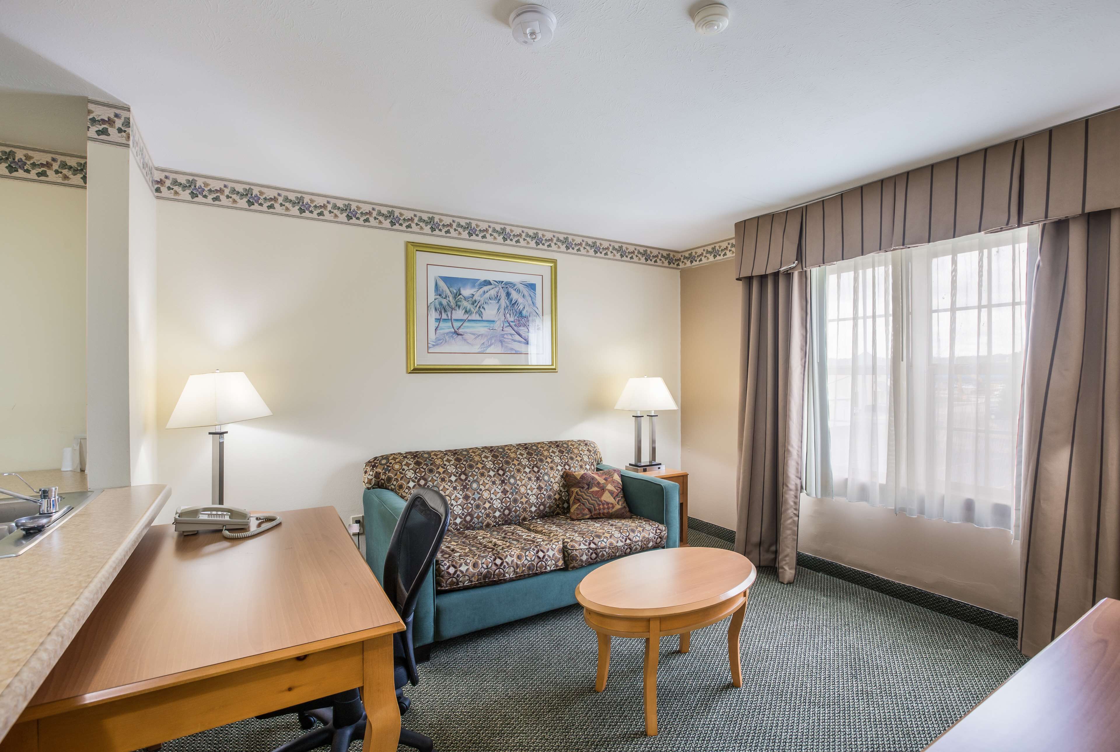 Une chambre d’hôtel avec un canapé, une petite table ronde, deux lampes, un tableau encadré au mur et une fenêtre avec des rideaux.
