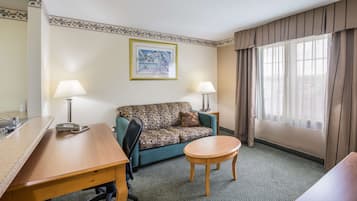 Une chambre d’hôtel avec un canapé, une petite table ronde, deux lampes, un tableau encadré au mur et une fenêtre avec des rideaux.