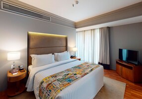 A hotel room with a large bed, two bedside tables with lamps, a TV on a wooden cabinet, and a window with curtains.