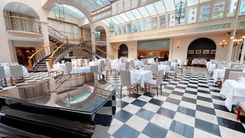 A spacious hotel lobby with a checkered floor, multiple dining areas, a grand staircase, and a high ceiling with skylights.