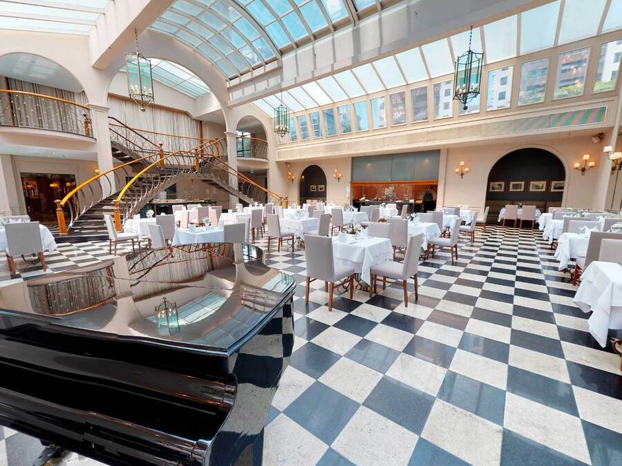 A spacious hotel lobby with a checkered floor, multiple dining areas, a grand staircase, and a high ceiling with skylights.