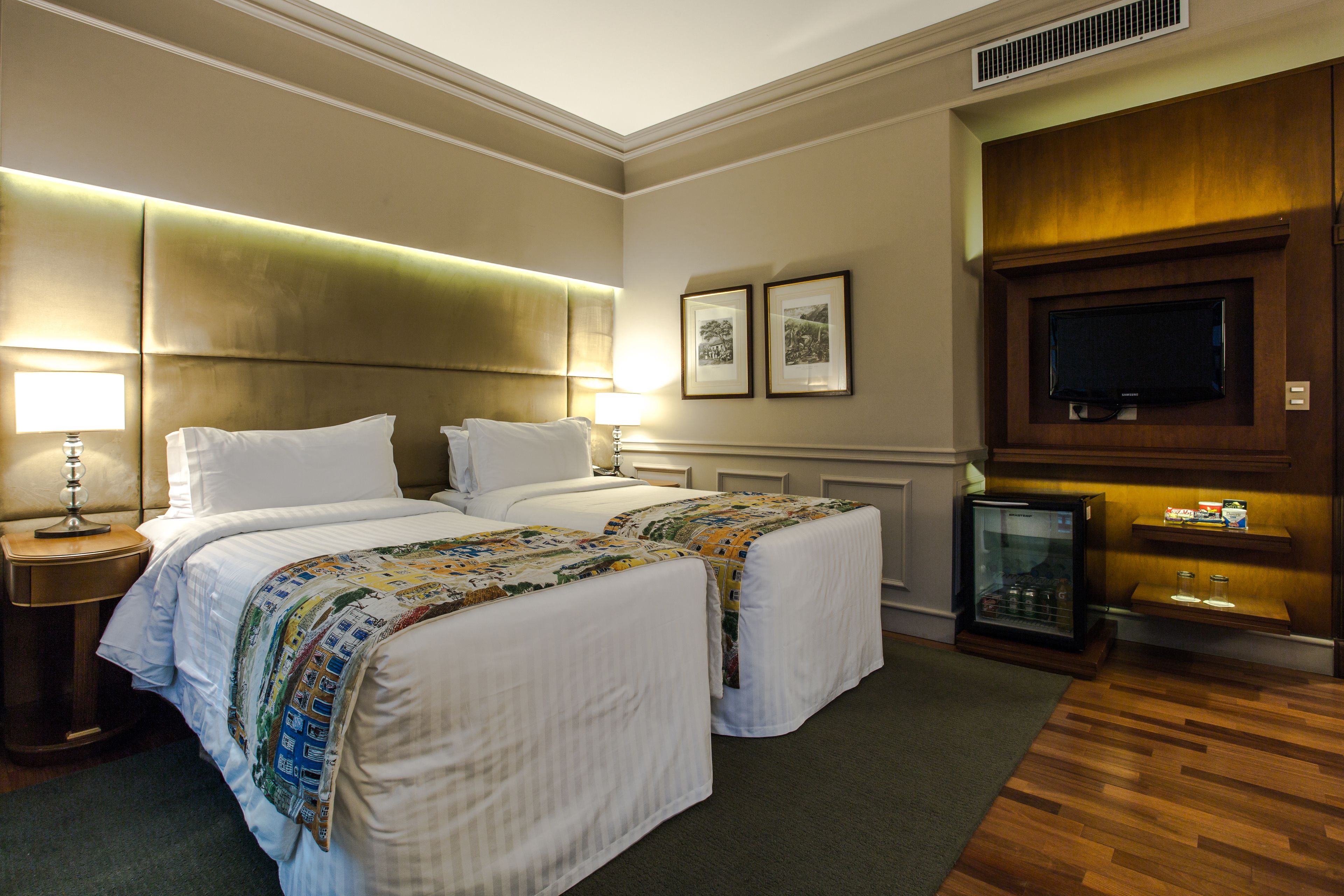 A hotel room with two beds, a television, a minibar, and framed pictures on the wall.