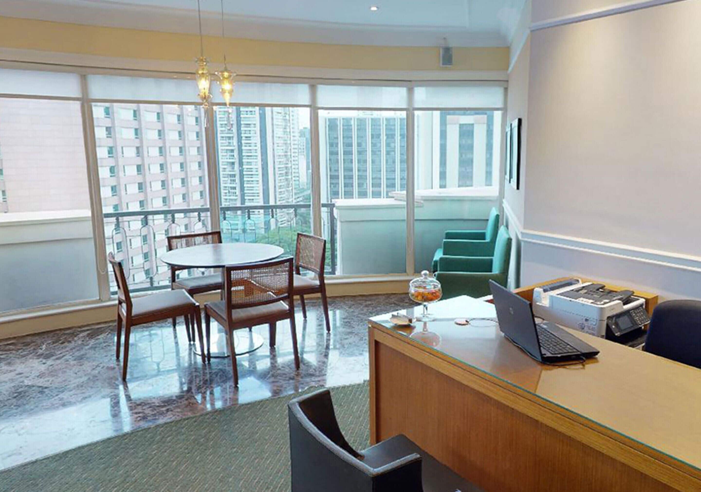 A modern office space with a round table, chairs, a desk with a laptop, and a view of tall buildings through large windows.