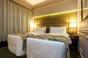 A hotel room with two beds, a headboard with built-in lighting, a bedside table with a lamp, and a large window with curtains.