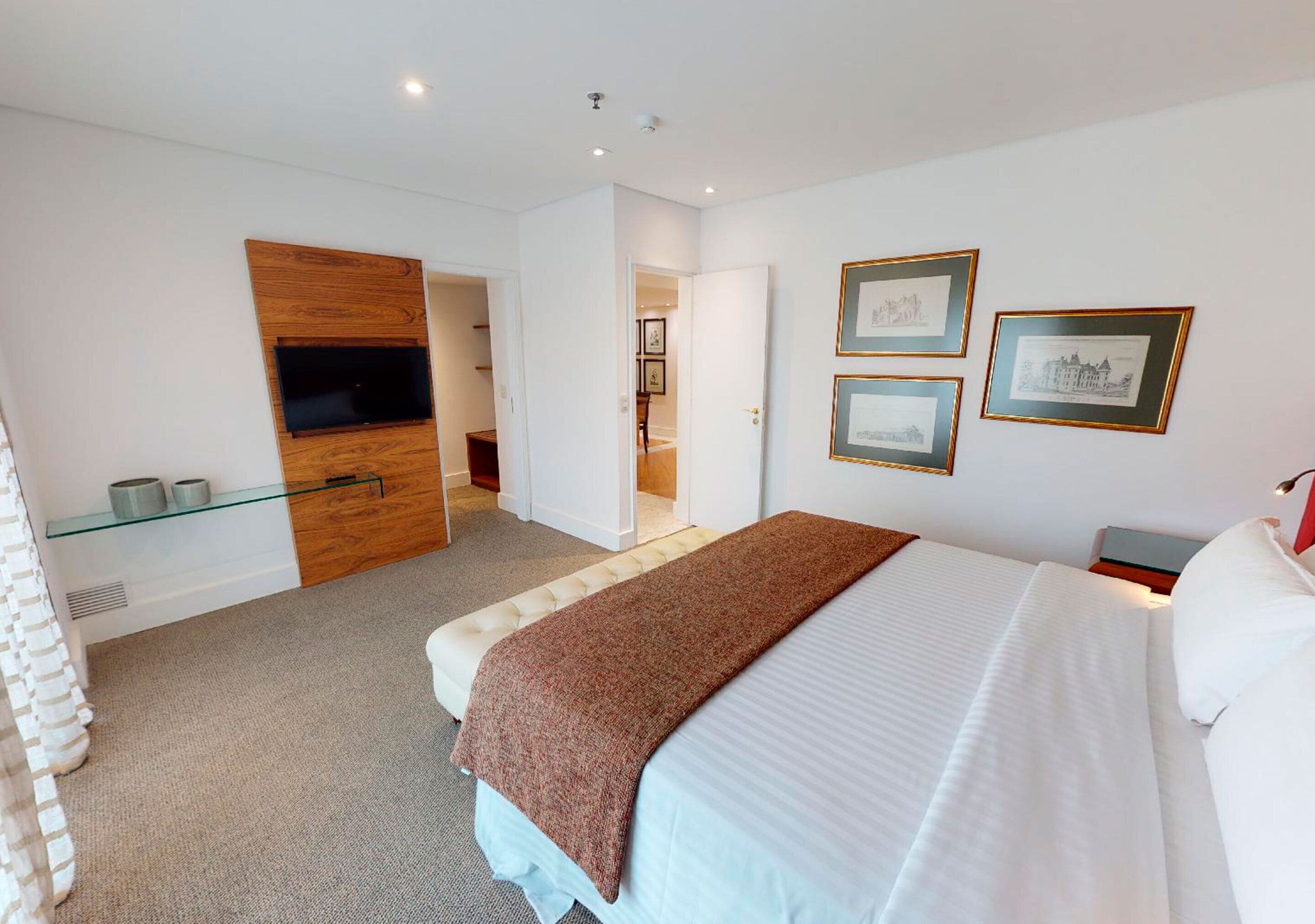 Een moderne hotelkamer met een bed, een aan een houten paneel bevestigde televisie, een glazen plank met toiletartikelen en ingelijste foto’s aan de muur.