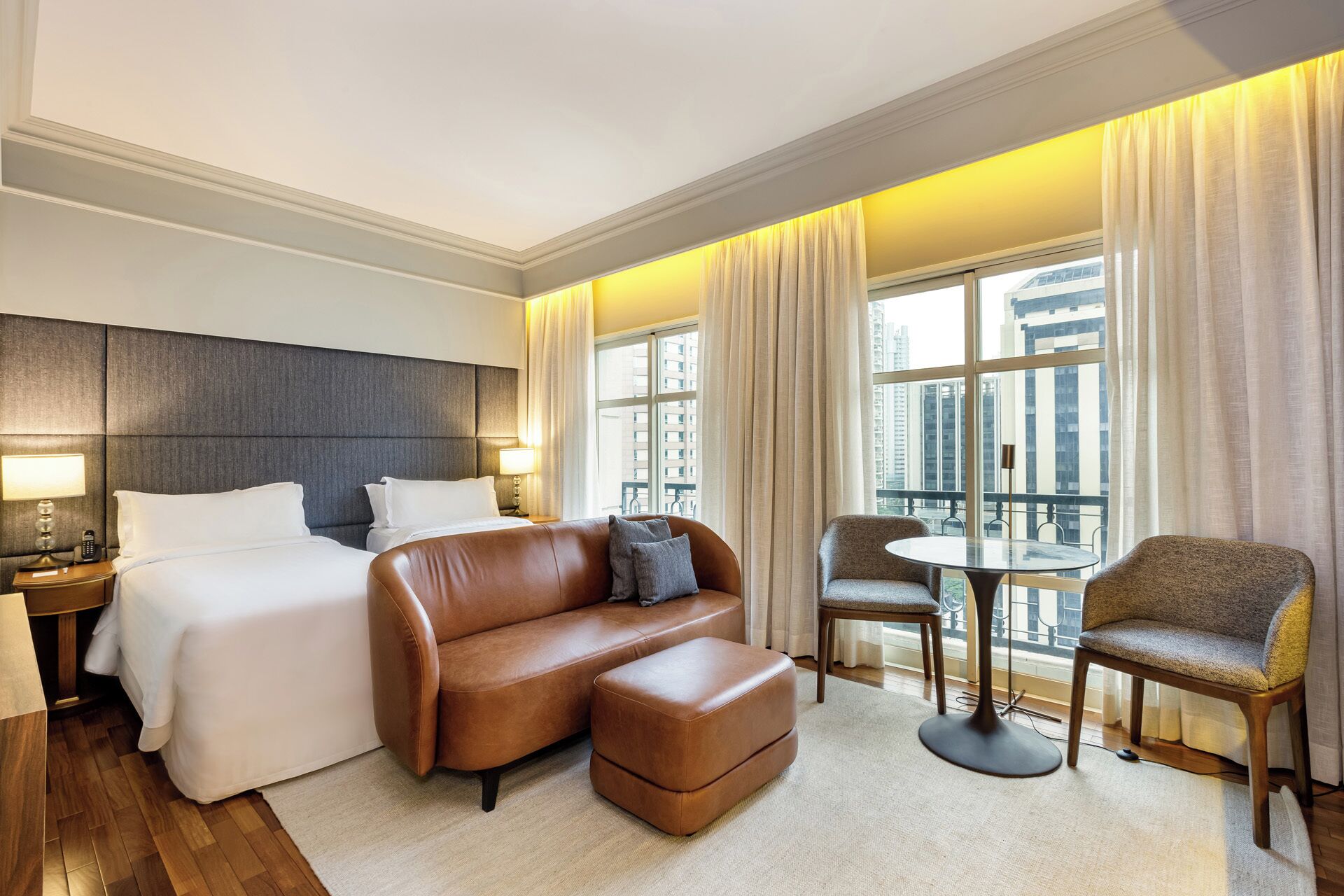 Una habitación de hotel con una cama, un sofá de cuero marrón, una mesita con sillas y un ventanal con cortinas.