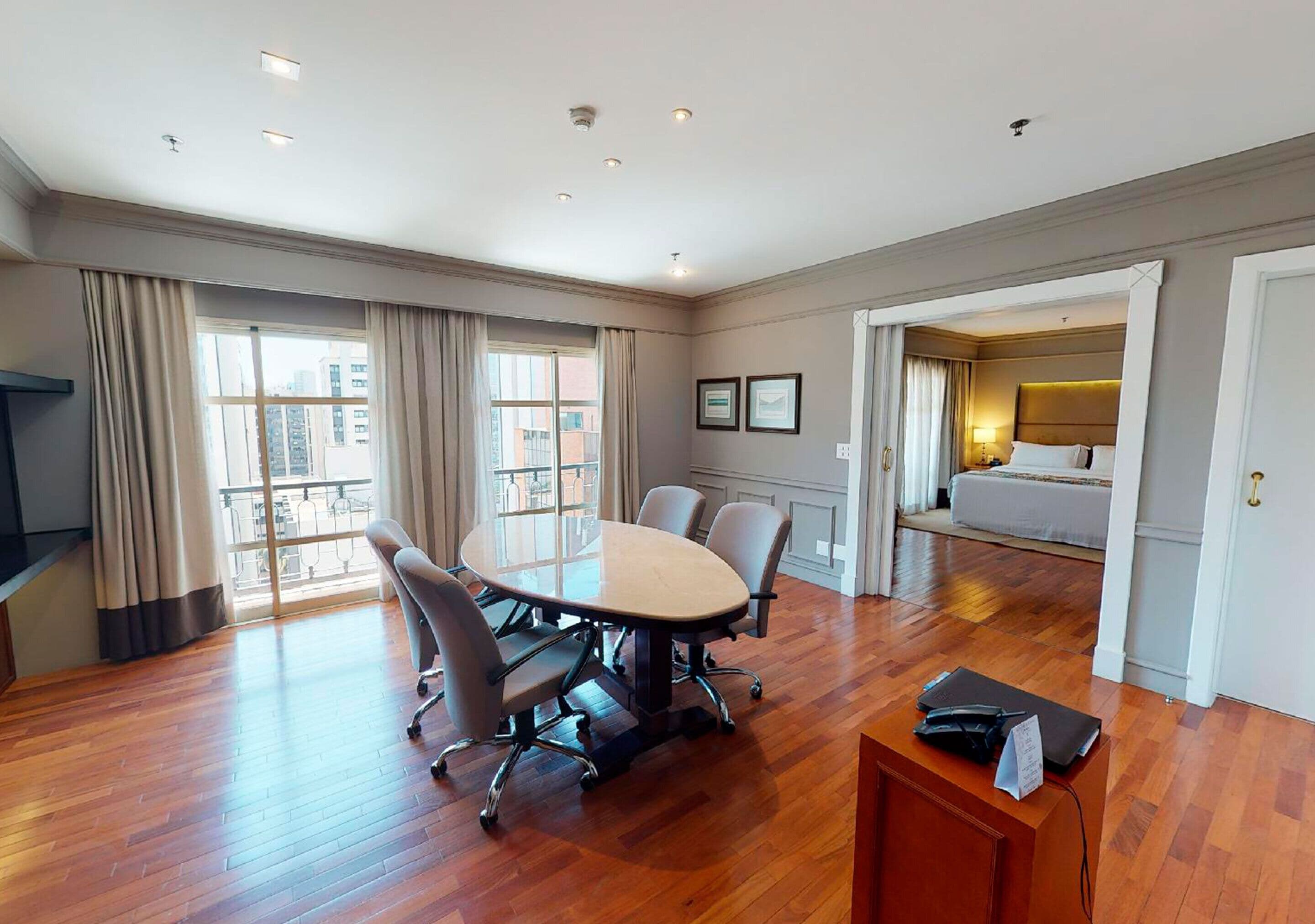 Quarto de hotel com piso de madeira, mesa de conferência com cadeiras, uma cama e banheiro visível por uma porta aberta.