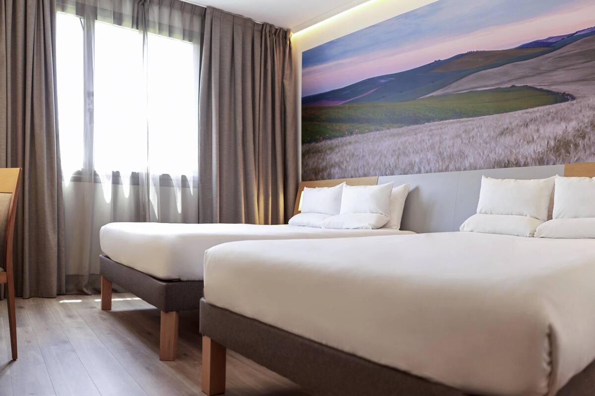 A hotel room with two beds, a large window with curtains, and a mural of a landscape.