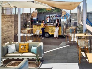 An outdoor seating area with a bar, tables, and chairs, and a food truck in the background. - Novotel Sevilla (Seville)