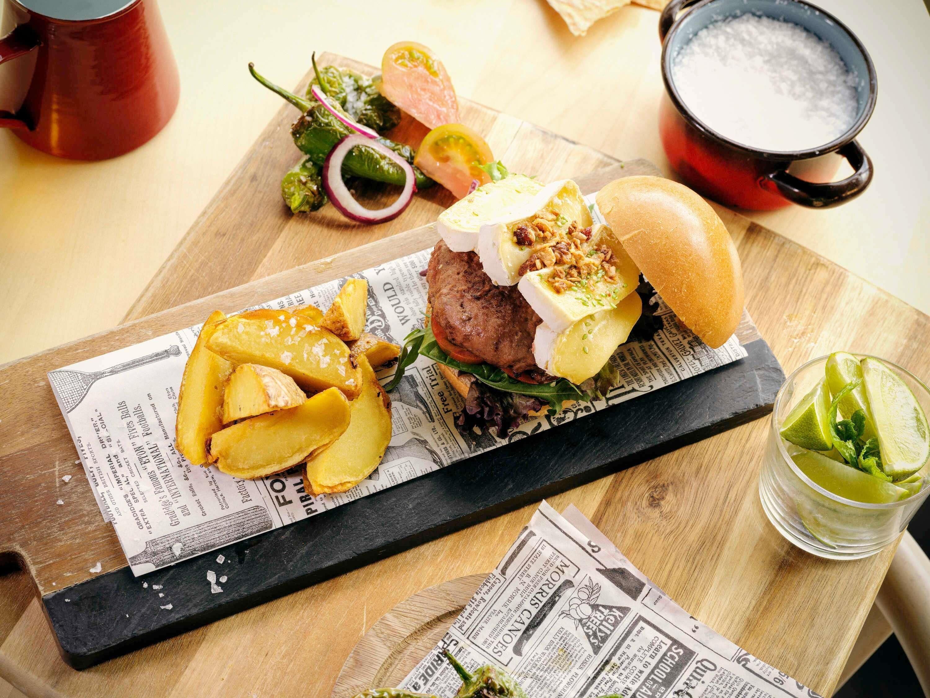Comida con una hamburguesa, papas fritas y una bebida sobre una mesa de madera, con periódicos debajo.