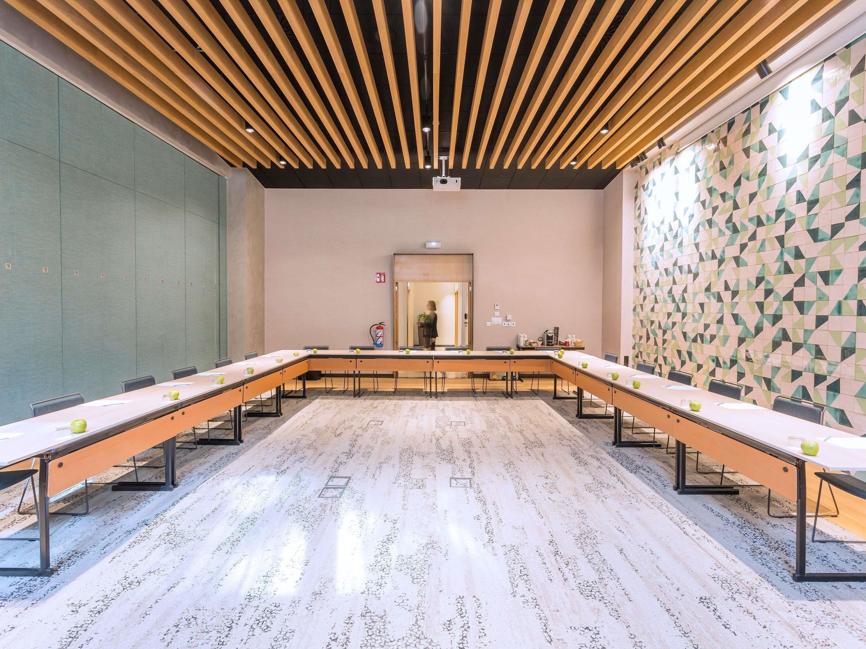 Un salón de conferencias con una larga mesa, sillas, techo con vigas de madera y un papel tapiz decorativo, además de un espejo grande.