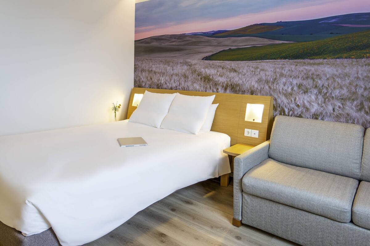 A hotel room with a bed, a nightstand, a lamp, a sofa, and a wall mural of a landscape.