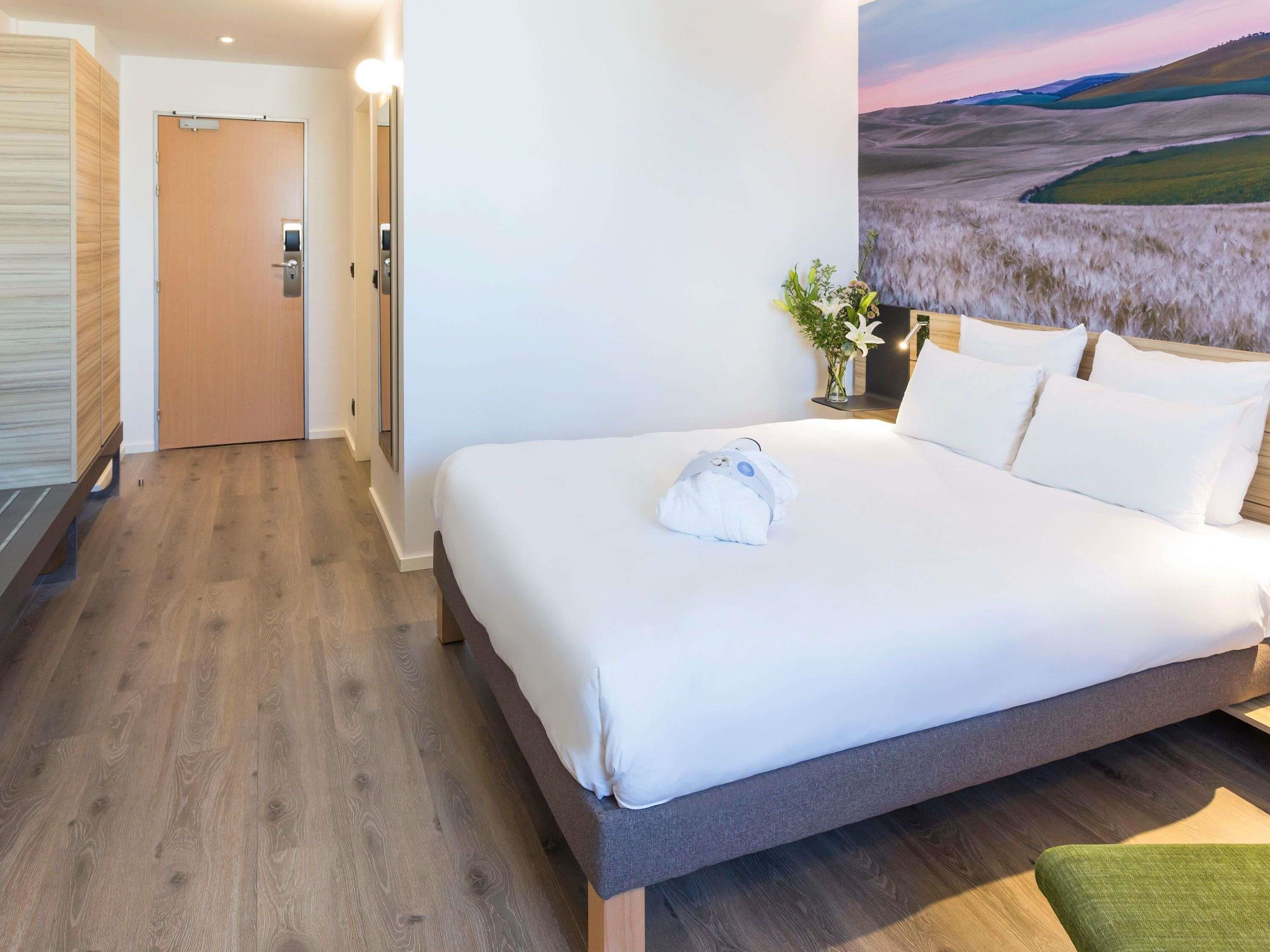 A hotel room with a large bed, a wooden floor, a door, and a wall with a landscape painting.