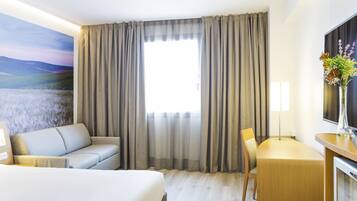 Habitación de hotel con una cama grande, un sofá, un escritorio y una silla. Se ve una ventana con cortinas, un cuadro en la pared y un florero con flores sobre el escritorio.