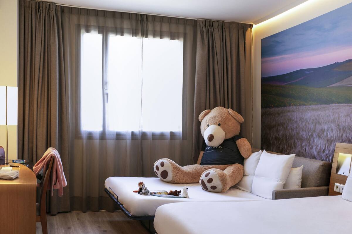 A hotel room with two beds, a large teddy bear on one bed, a painting of a landscape on the wall, and a desk with a lamp.