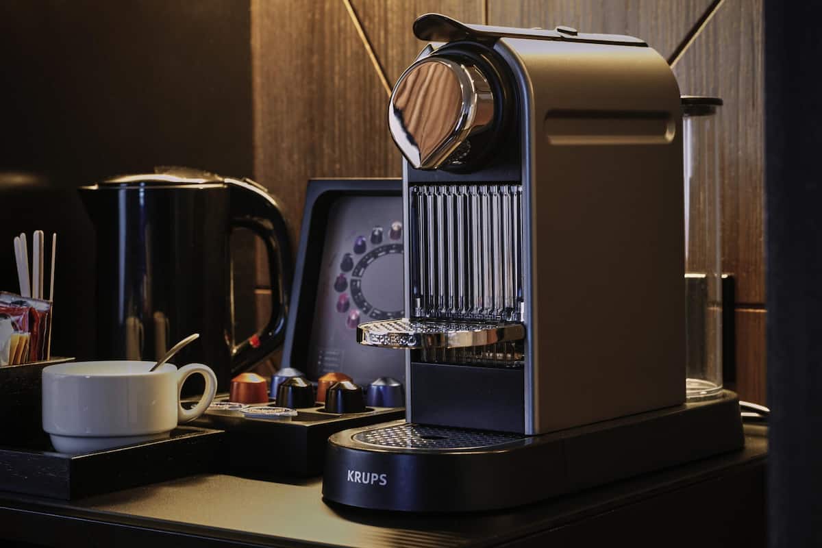 A Krups coffee maker on a countertop with a white mug, a coffee pot, and a coffee grinder.