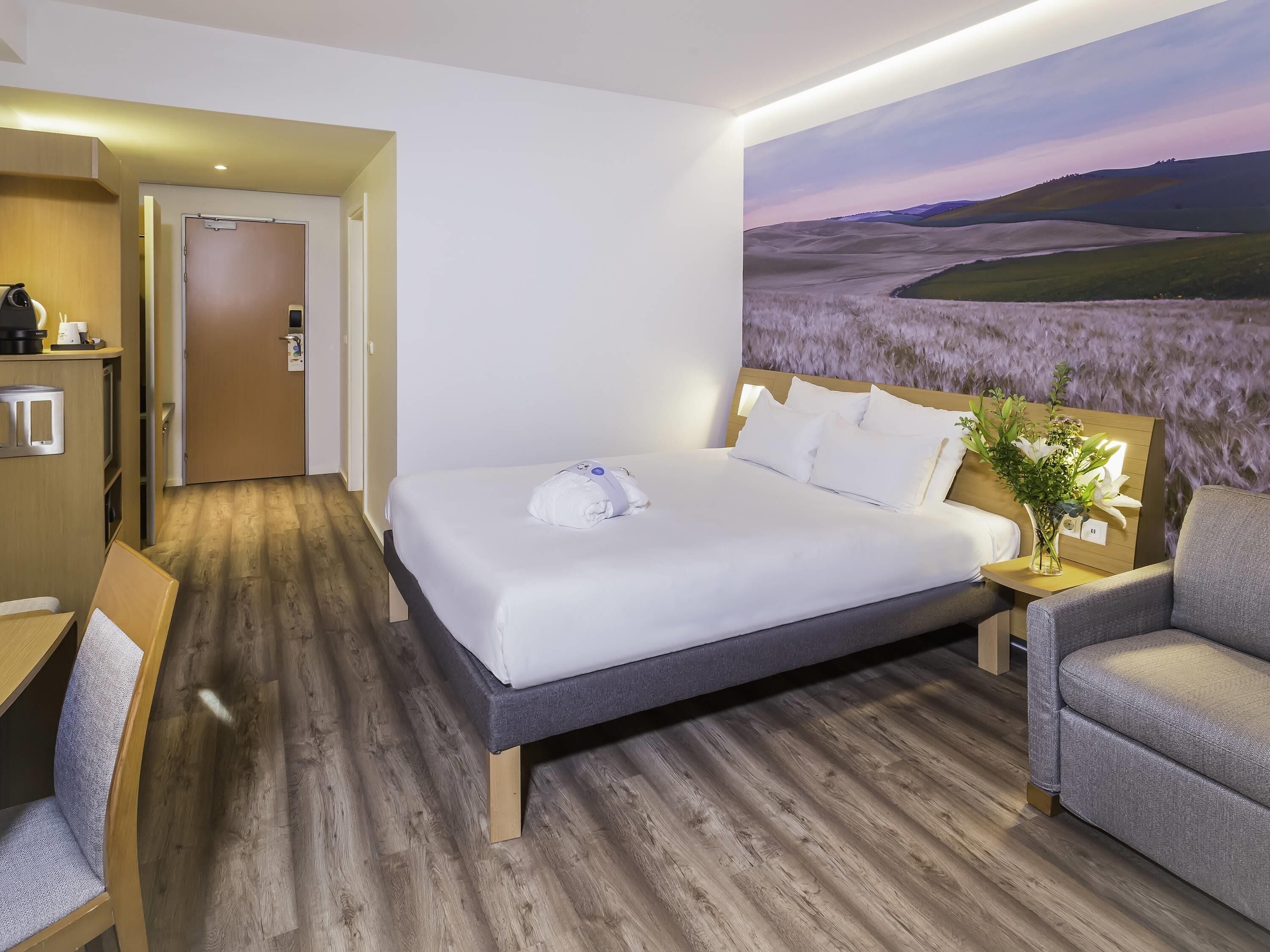 A hotel room with a bed, a desk, a chair, a nightstand, and a wall-mounted TV.