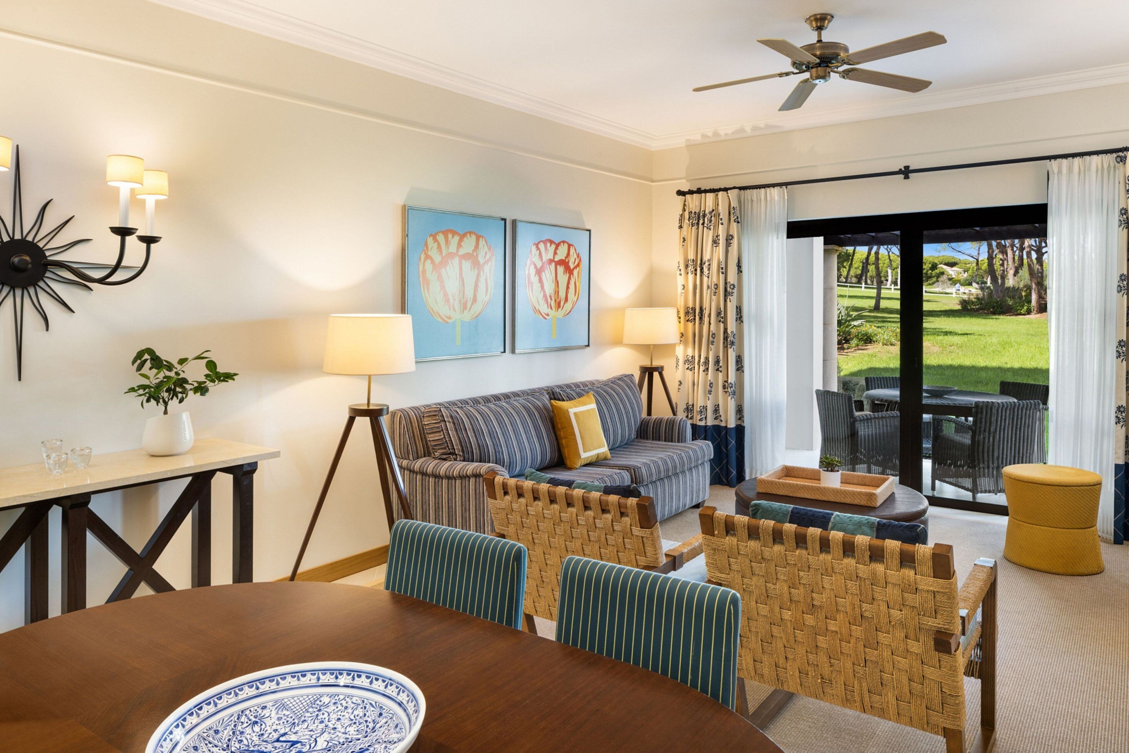 A dining area with a round table, chairs, and a decorative plate. There is a sofa with a patterned cushion, a floor lamp, and a painting on the wall.