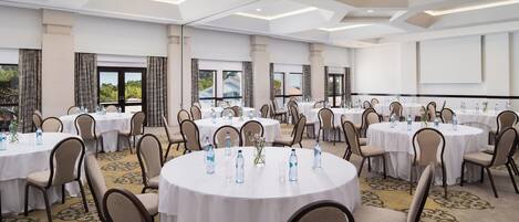 A conference room with round tables, chairs, and water bottles, featuring a carpeted floor and large windows.