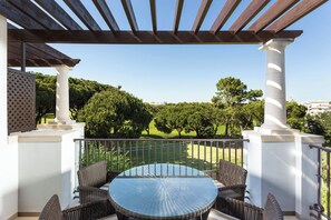 A covered outdoor patio with a round glass table and wicker chairs, overlooking a green landscape.