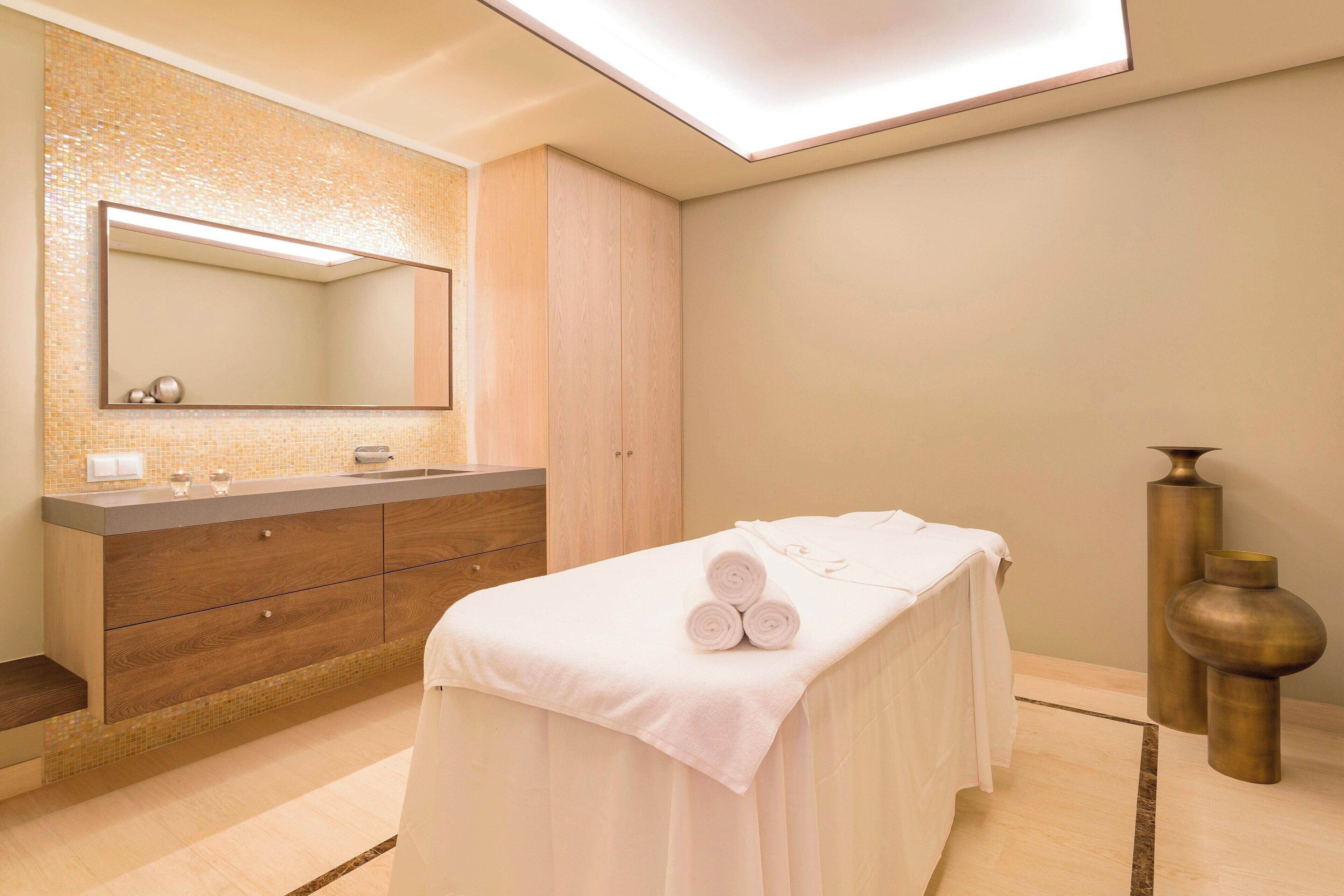 A modern spa room with a wooden vanity, a large mirror, a massage table with towels, and two decorative vases.