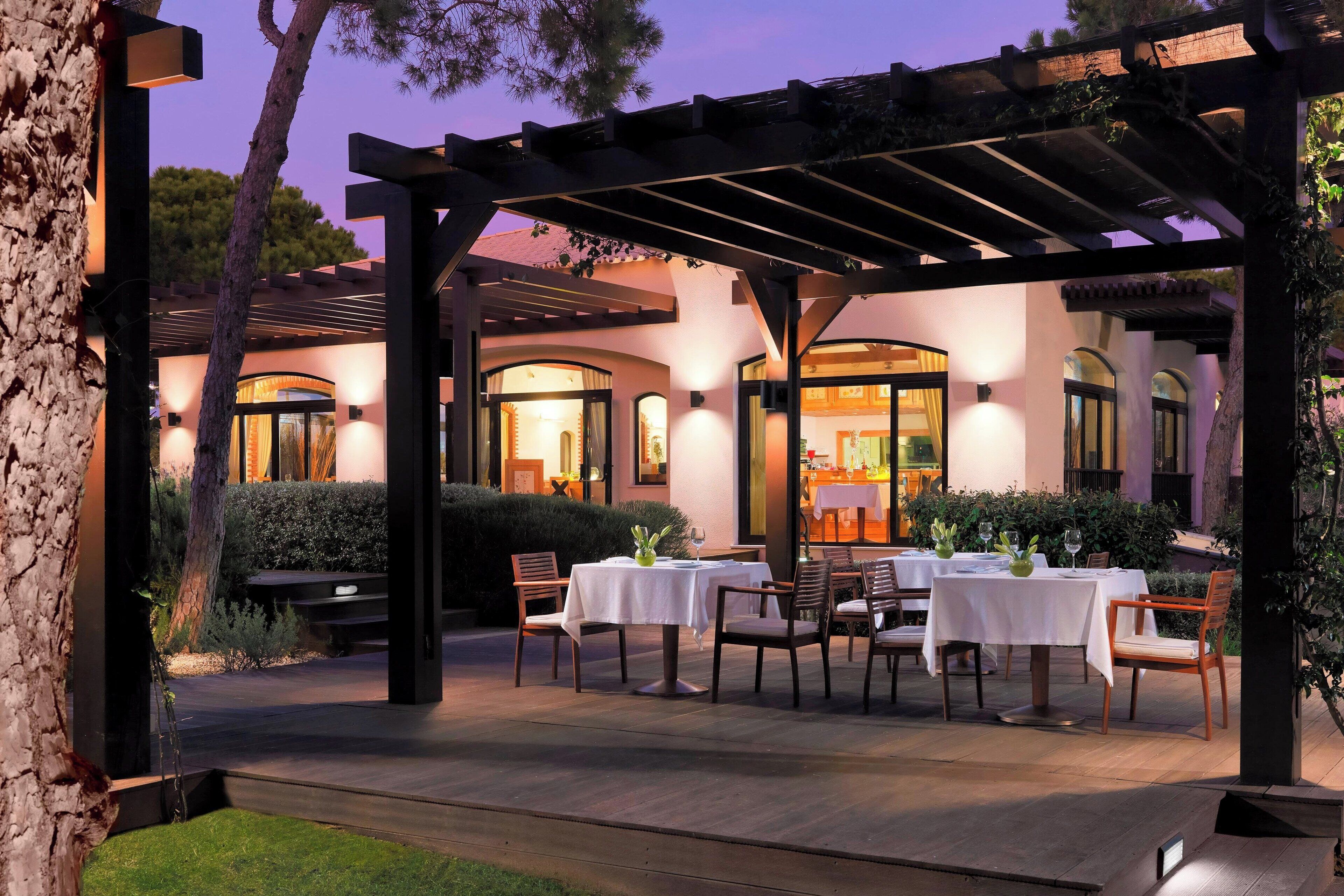 An outdoor dining area with tables and chairs under a pergola, surrounded by a well-maintained garden and a building with arched windows.