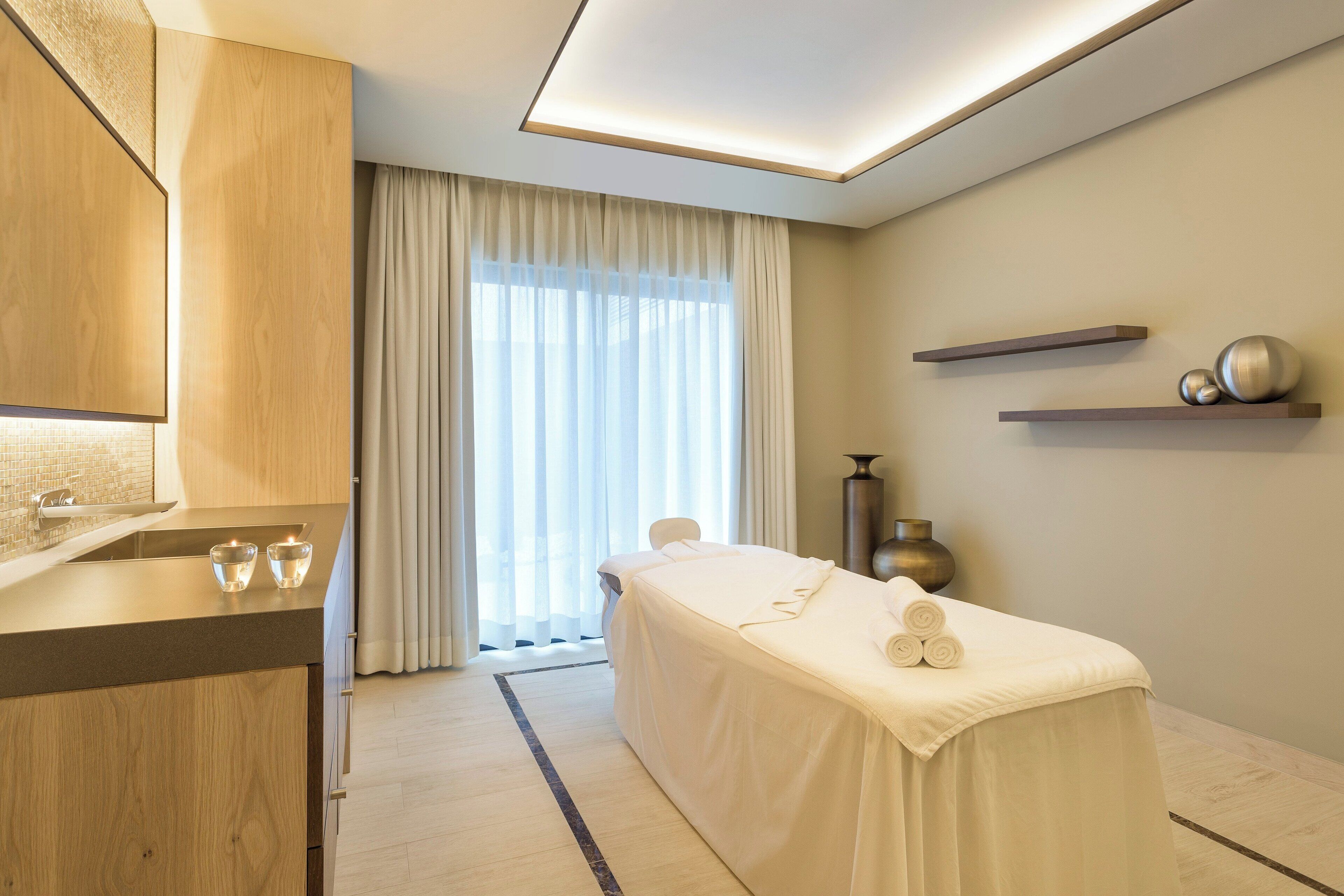 A modern spa room with a massage table, a wooden cabinet, and a large window with sheer curtains.