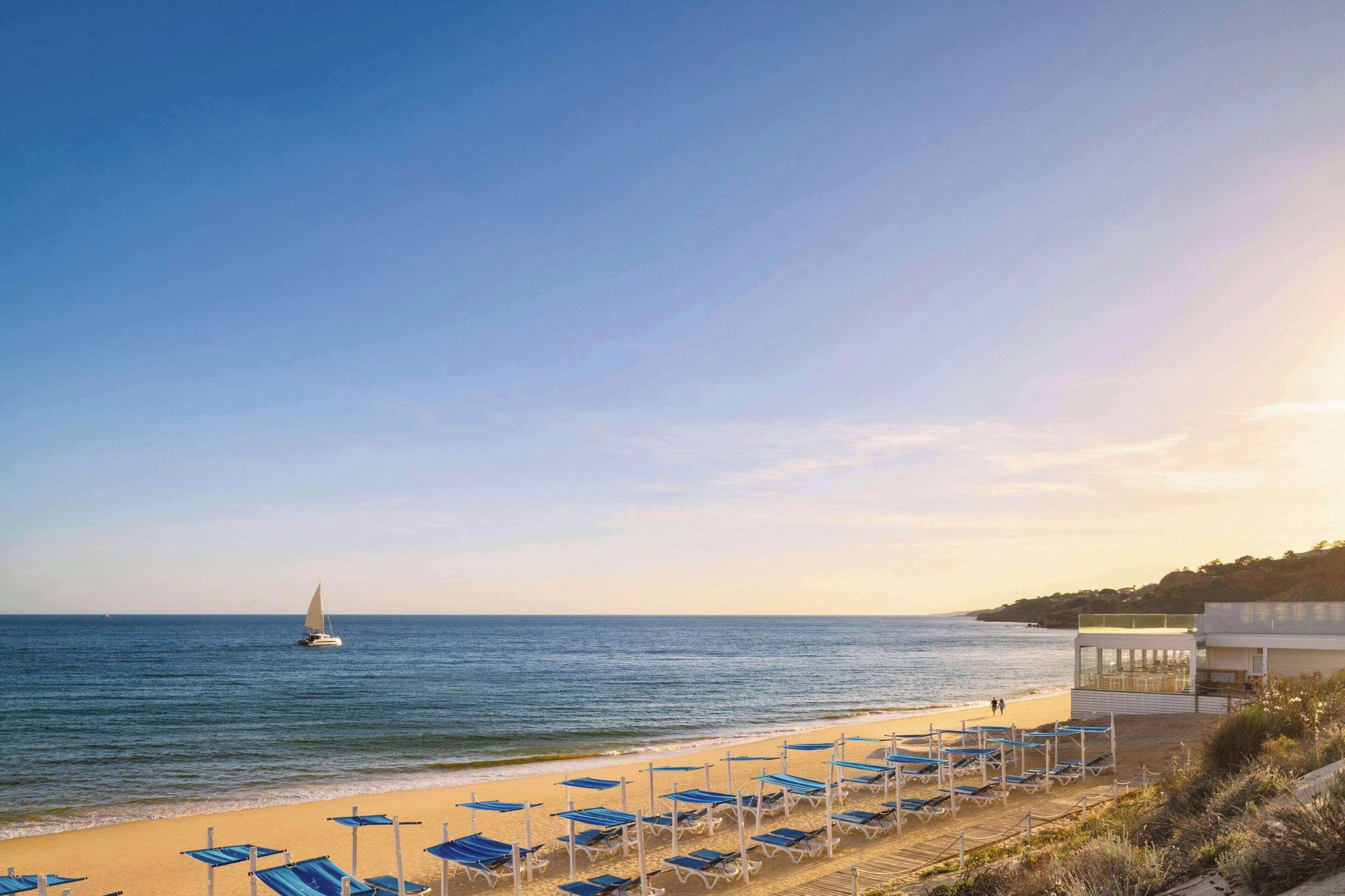 Une plage bordée de rangées de chaises longues bleues, et un voilier sur l’eau.