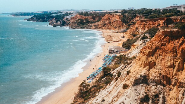 Une plage côtière bordée de falaises rocheuses, avec une étendue de sable et une rangée de parasols bleus.