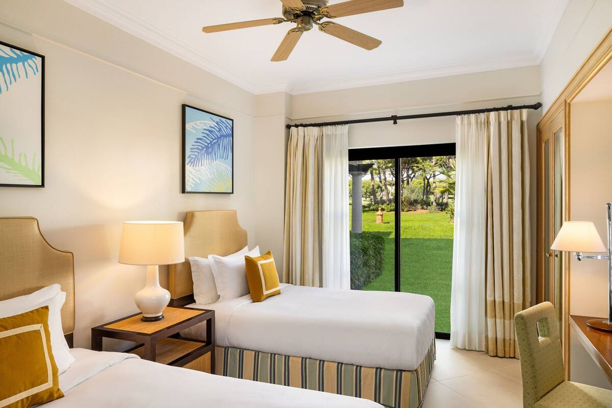 A hotel room with two beds, a ceiling fan, a view of a lawn through a sliding door, and artwork on the walls.