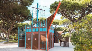A wooden play structure with a ship-themed design, including a mast and anchor, surrounded by trees and a sandy ground.