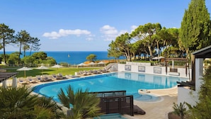 Une piscine entourée d’arbres, avec des chaises longues et des parasols, offrant une vue sur la mer.