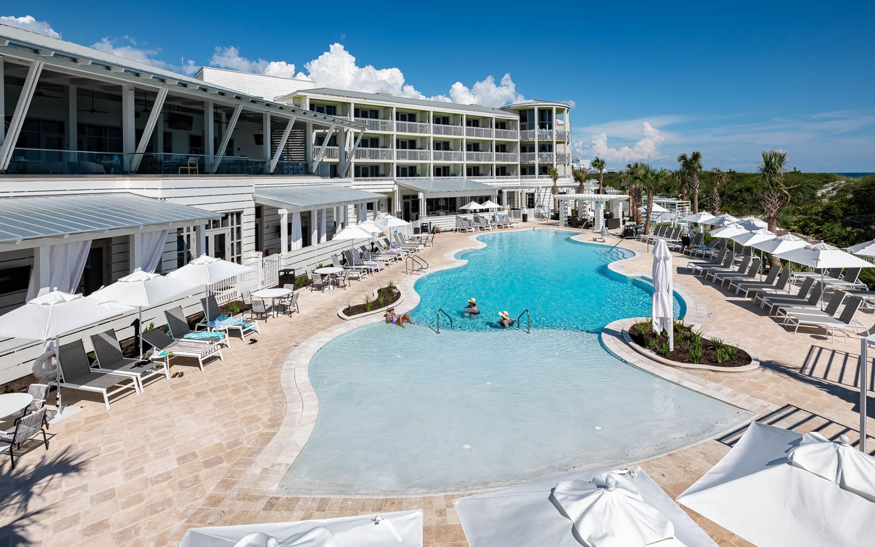 Un resort con una piscina grande, sillones de sol, sombrillas y un edificio de varios pisos con balcones.