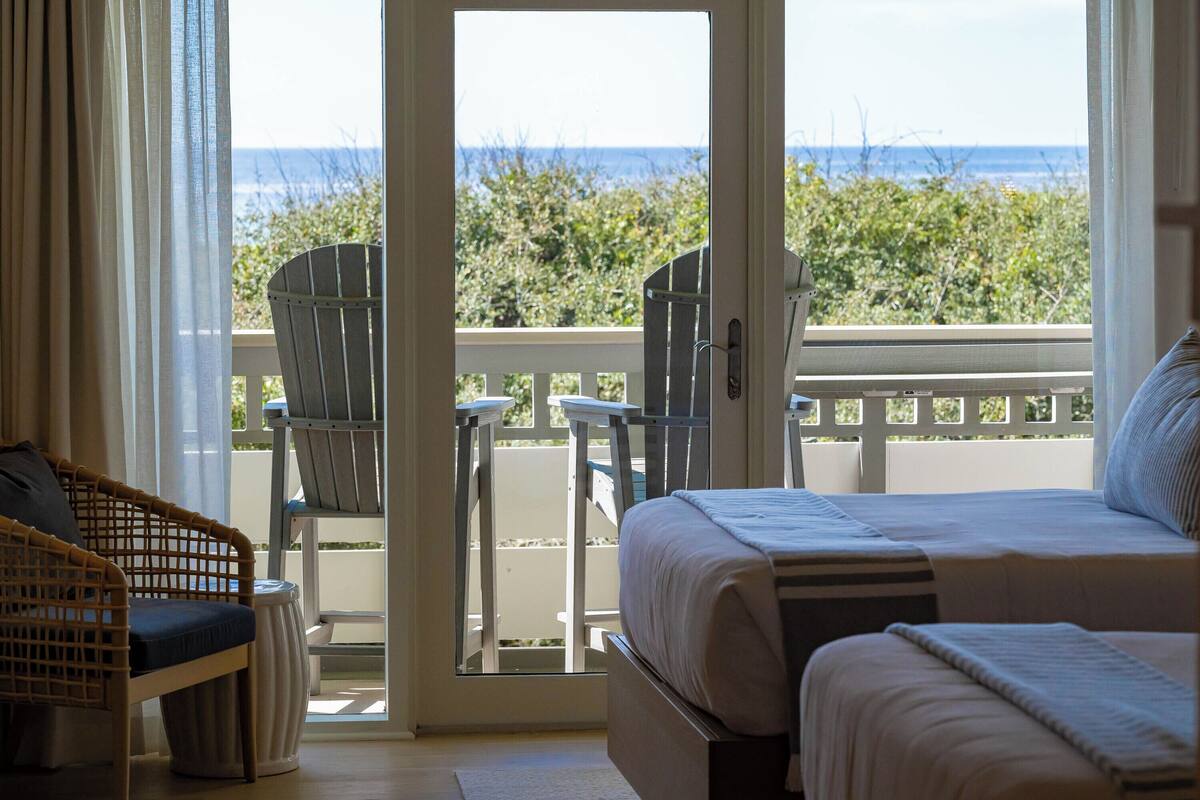 A hotel room with a bed, a wicker chair, a small table, and a view of the beach and ocean.