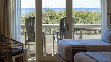 A hotel room with a bed, a wicker chair, a small table, and a view of the beach and ocean.