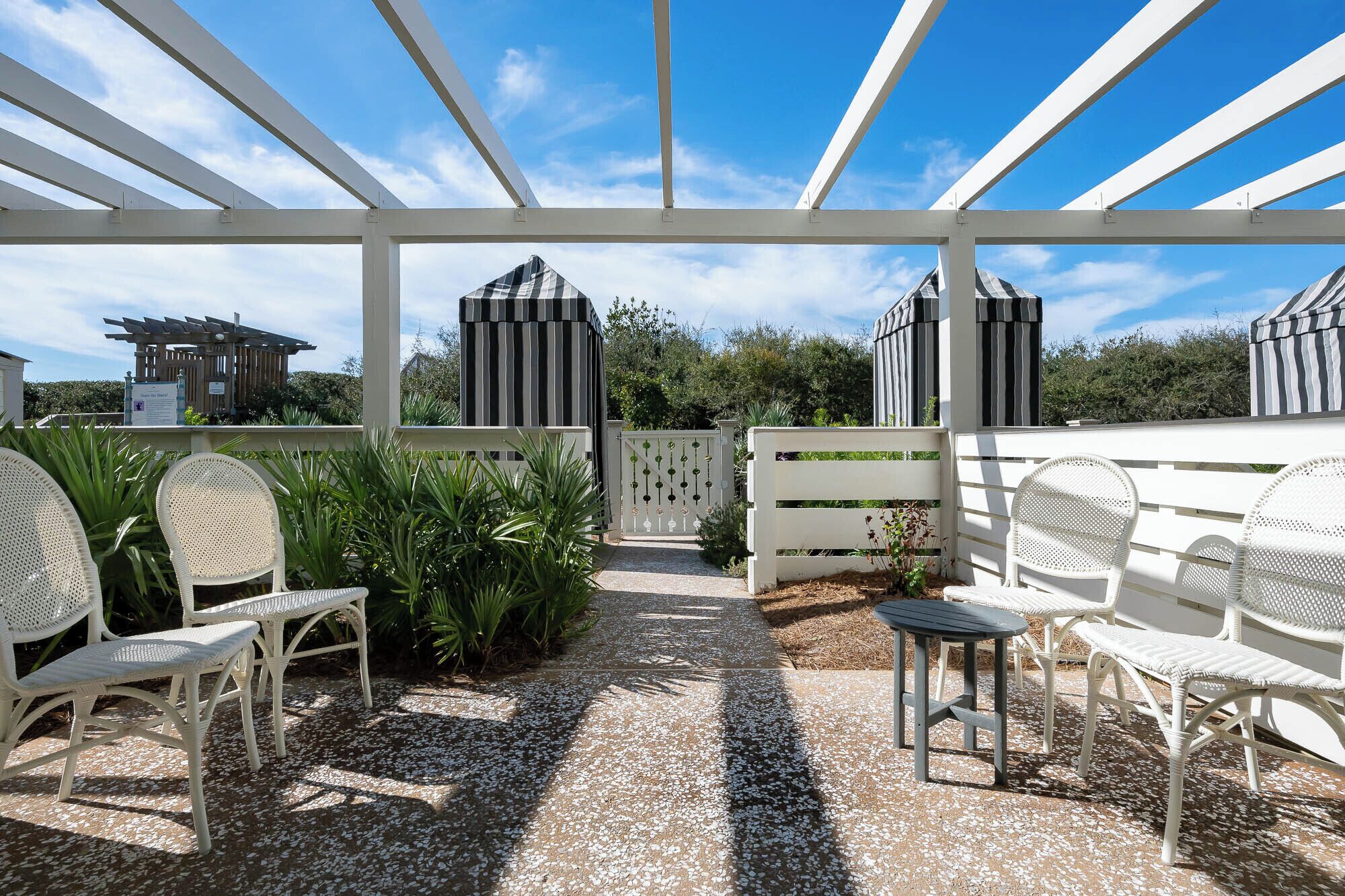 An outdoor patio area with white wicker furniture, a gravel path, and a view of a building with striped awnings.