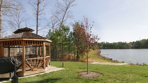 Un pabellón de madera con techo, una parrilla y un banco, ubicado junto a un lago con árboles y un espacio cubierto de césped.