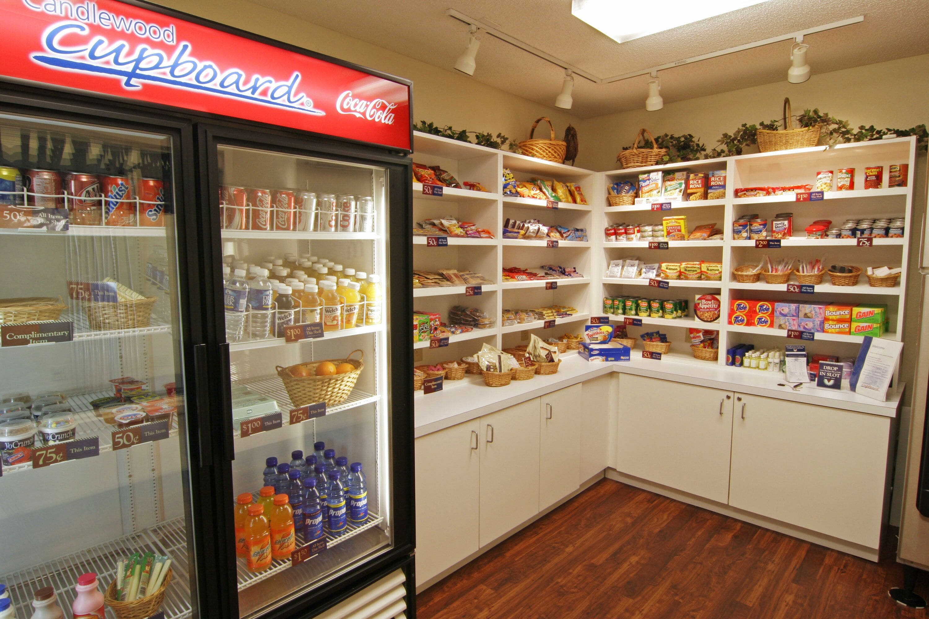 Une petite cuisine équipée d’un réfrigérateur rempli de boissons, d’étagères avec des en-cas et d’une enseigne indiquant « Cupboard Coca-Cola ».