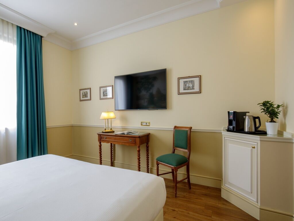 Una camera d'albergo con un letto, una scrivania con una lampada, una sedia, una televisione, quadri appesi alle pareti e una macchina per il caffè.