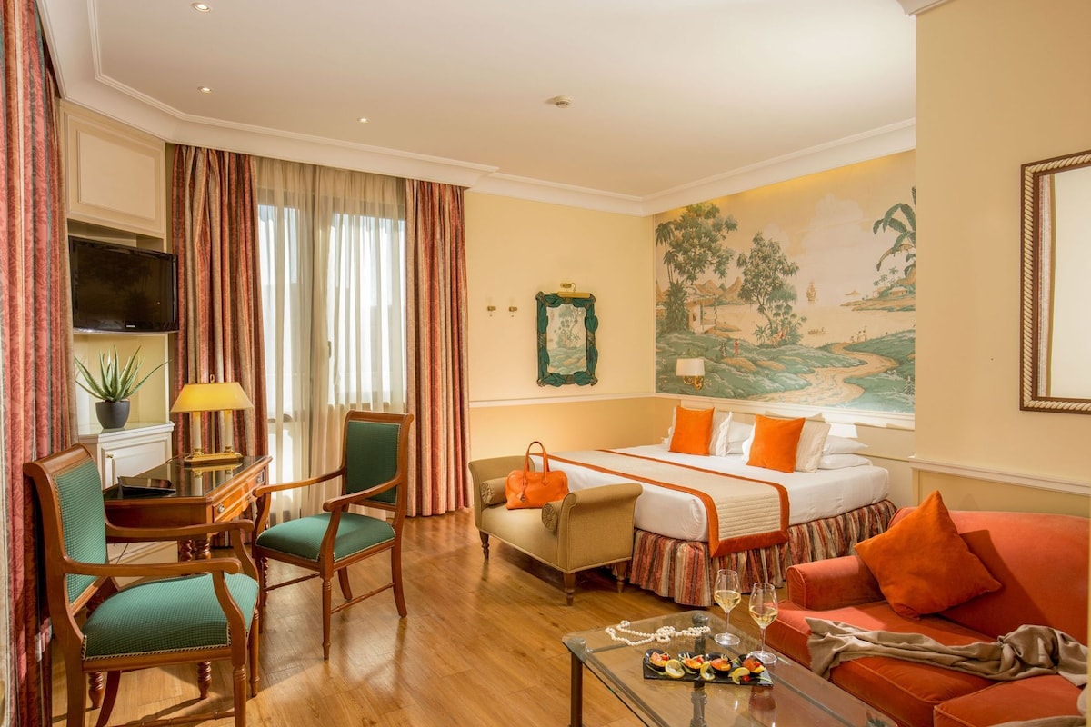 Habitación de hotel con cama, dos sillas, una mesa con lámpara, un televisor y un cuadro en la pared.