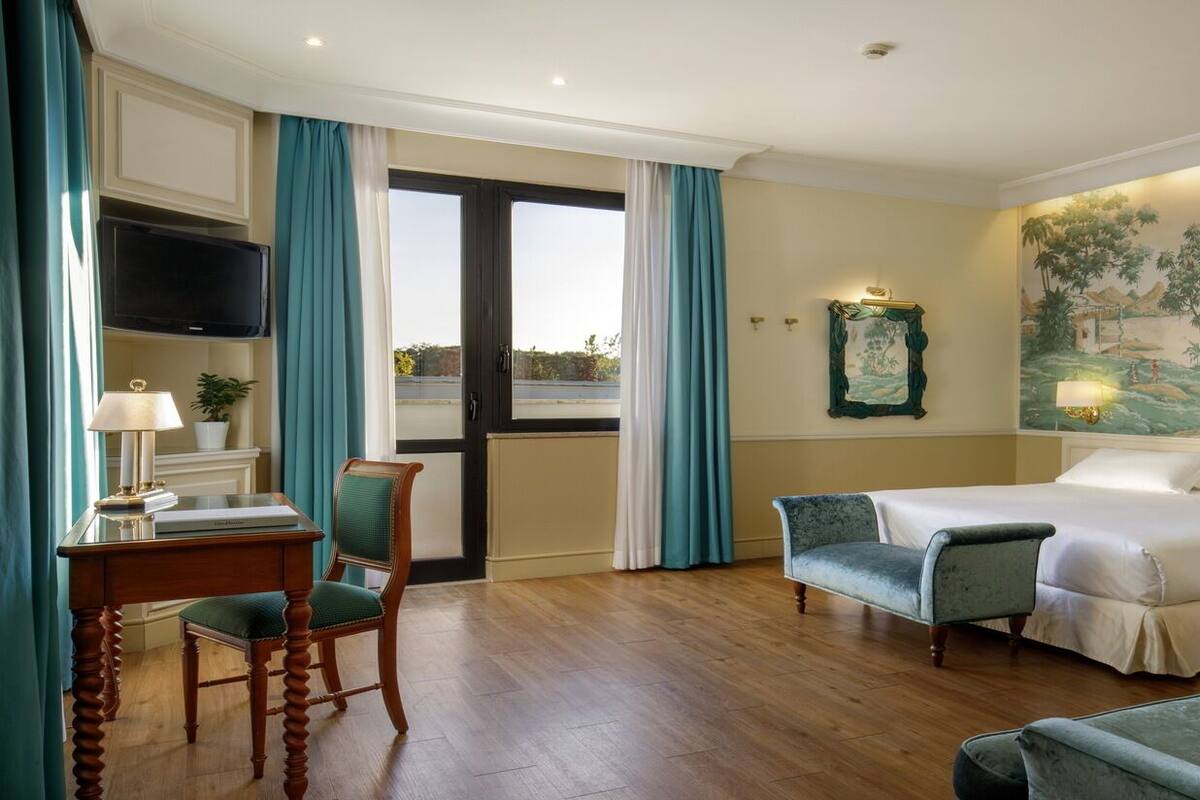 A hotel room with a bed, a desk with a lamp, a chair, a television, and a mural on the wall.