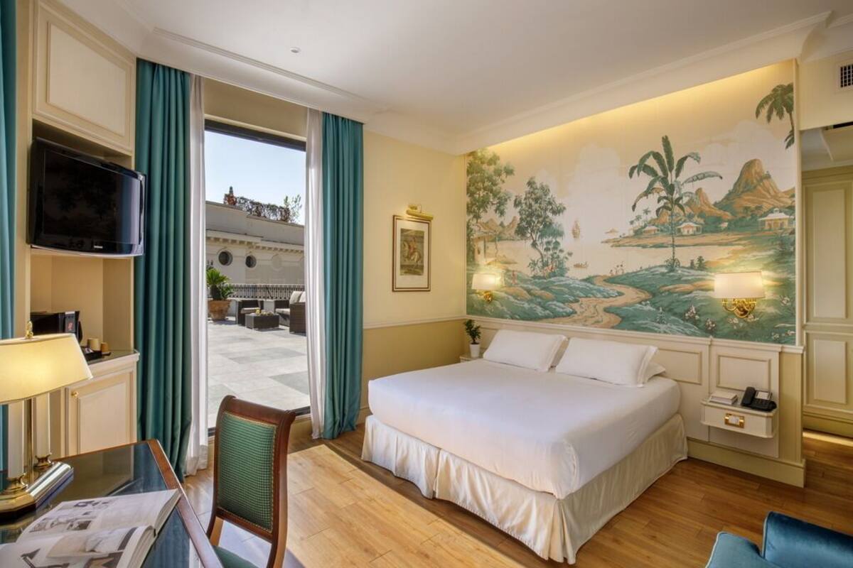 A hotel room with a bed, a desk with a lamp, a chair, a TV, and a mural on the wall.