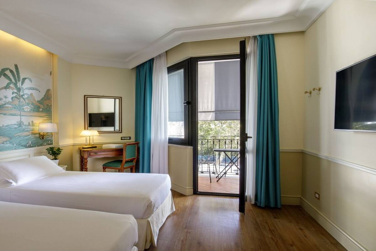 A hotel room with two beds, a desk with a chair, a balcony with a table and chairs, and a large TV on the wall.