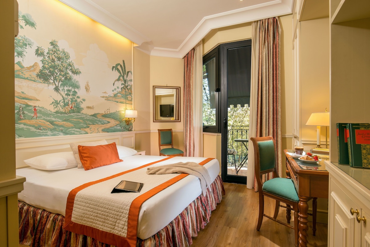 Habitación de hotel con cama, escritorio, silla, espejo, ventana con cortinas y un mural en la pared.